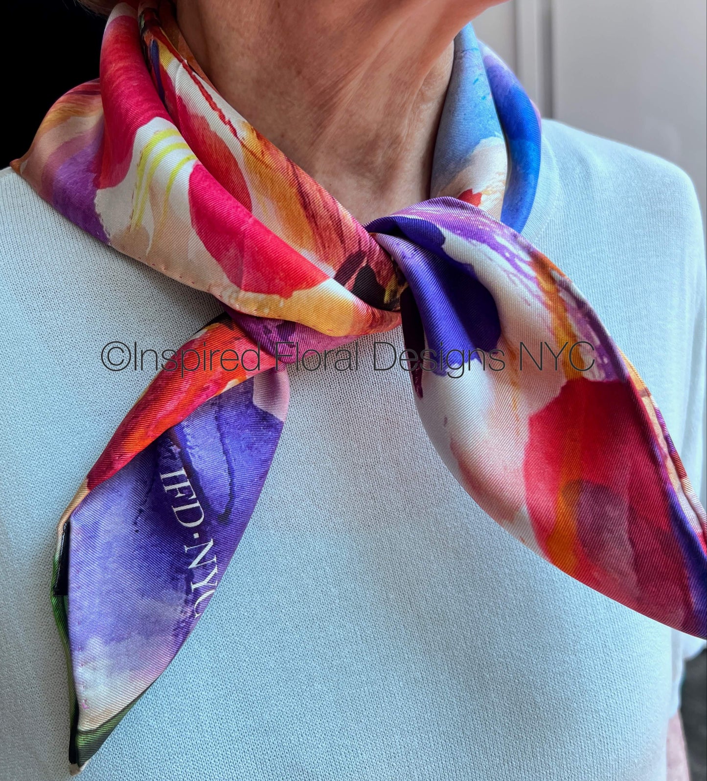 A close-up of a woman wearing a light blue top and a colorful floral silk scarf tied around her neck. The scarf features a vibrant abstract floral pattern by Inspired Floral Designs NYC LLC in pink, purple, blue, yellow, and white, with a visible copyright watermark '©Inspired Floral Designs NYC' across the center. Copyright