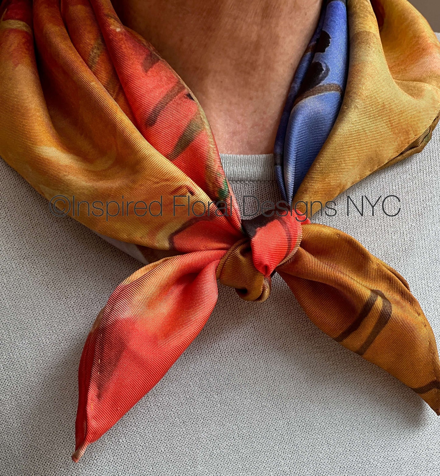 A close-up of a person wearing a patterned silk scarf by Inspired Floral Designs NYC LLC tied around the neck. The scarf features a warm gradient of orange, red, and yellow with dark brown abstract floral or leaf-like patterns, and a blue patterned section visible on the inside. Copyright
