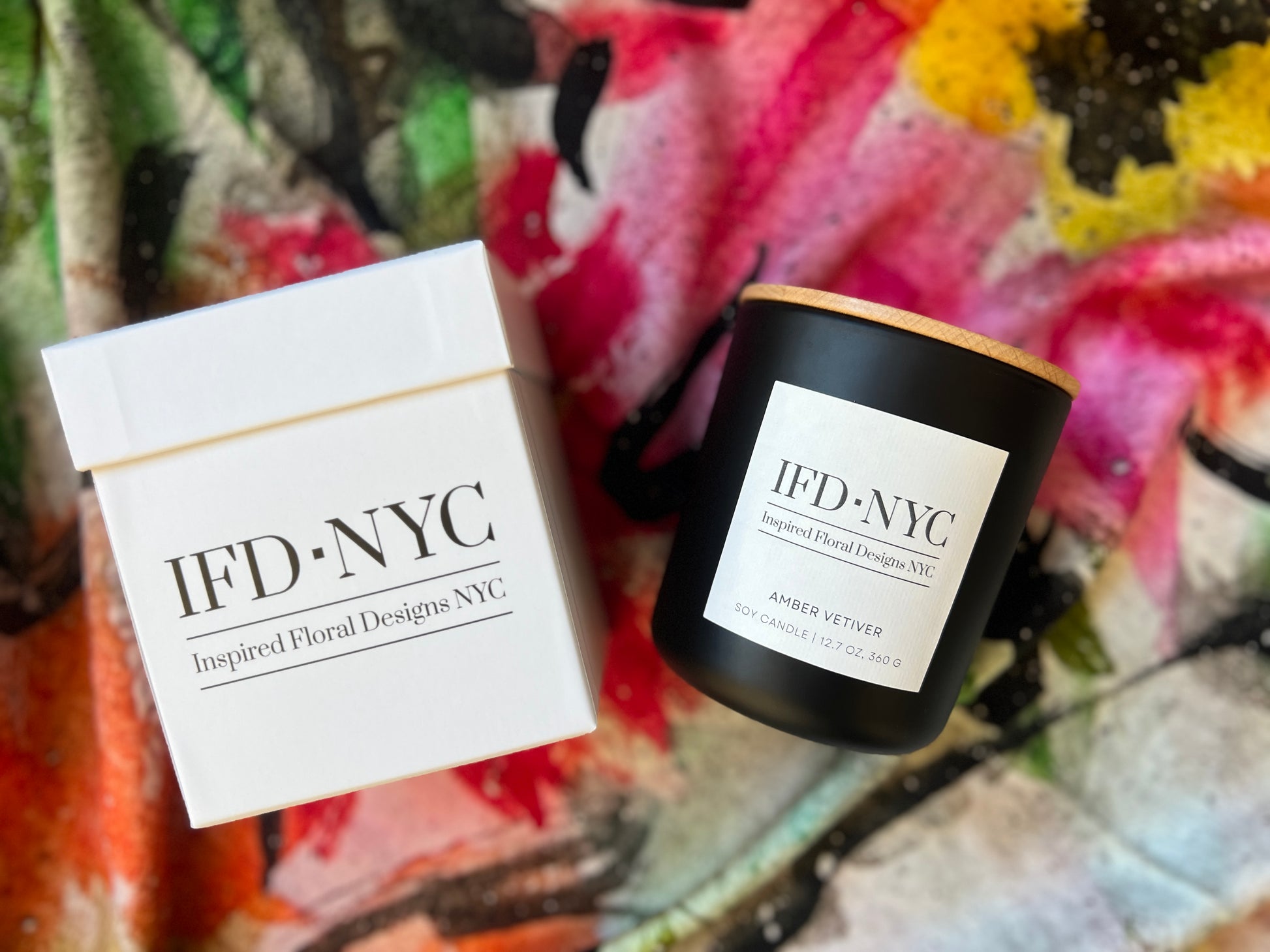 the inspired candle Black candle with wooden lid and white label on a colorful floral fabric background