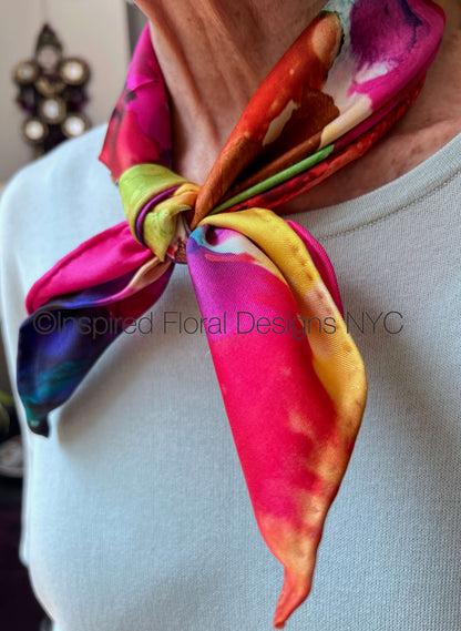 A close-up of a person wearing a vibrant silk scarf tied around the neck. The scarf features a multicolor floral and abstract pattern with prominent shades of pink, red, yellow, green, and blue by Inspired Floral Designs NYC LLC Copyright 