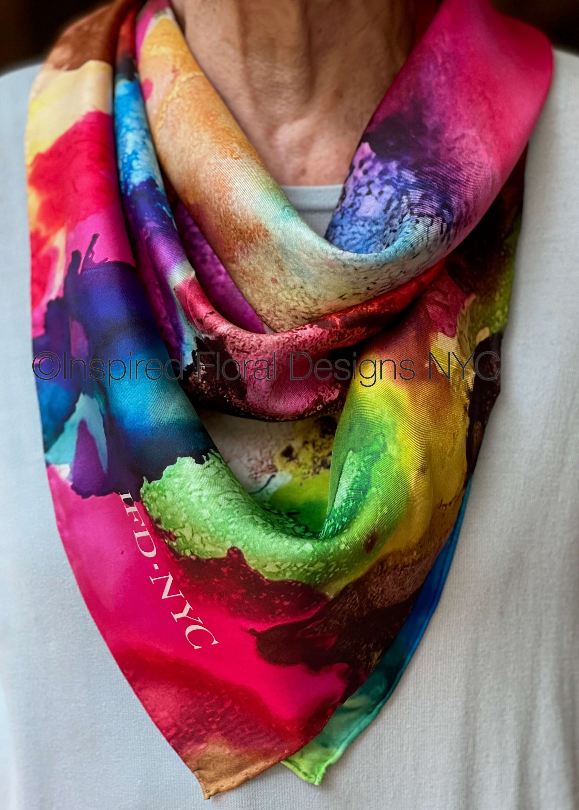 A close-up of a person wearing a vibrant, multicolored abstract watercolor print scarf. The scarf features a mix of bright pink, green, blue, yellow, and purple tones with a watercolor texture, draped around the neck over a light gray shirt. Inspired Floral Designs NYC copyright