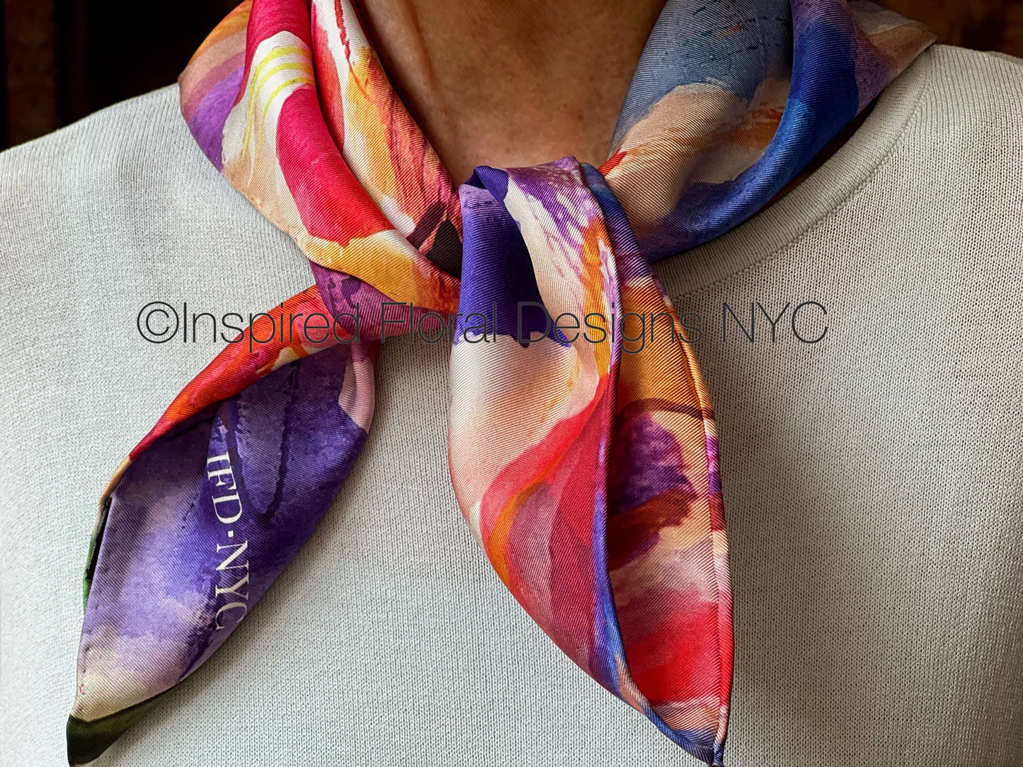 A close-up of a person wearing a square silk scarf tied around the neck. The scarf features an abstract watercolor pattern by Inspired Floral Designs NYC LLC with prominent shades of purple, red, orange, yellow, and blue. Copyright
