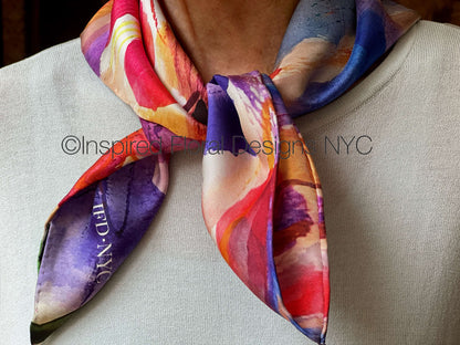 A close-up of a person wearing a square silk scarf tied around the neck. The scarf features an abstract watercolor pattern by Inspired Floral Designs NYC LLC with prominent shades of purple, red, orange, yellow, and blue. Copyright
