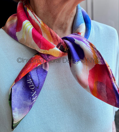 A close-up of a woman wearing a light blue top and a colorful floral silk scarf tied around her neck. The scarf features a vibrant abstract floral pattern by Inspired Floral Designs NYC LLC in pink, purple, blue, yellow, and white, with a visible copyright watermark '©Inspired Floral Designs NYC' across the center. Copyright