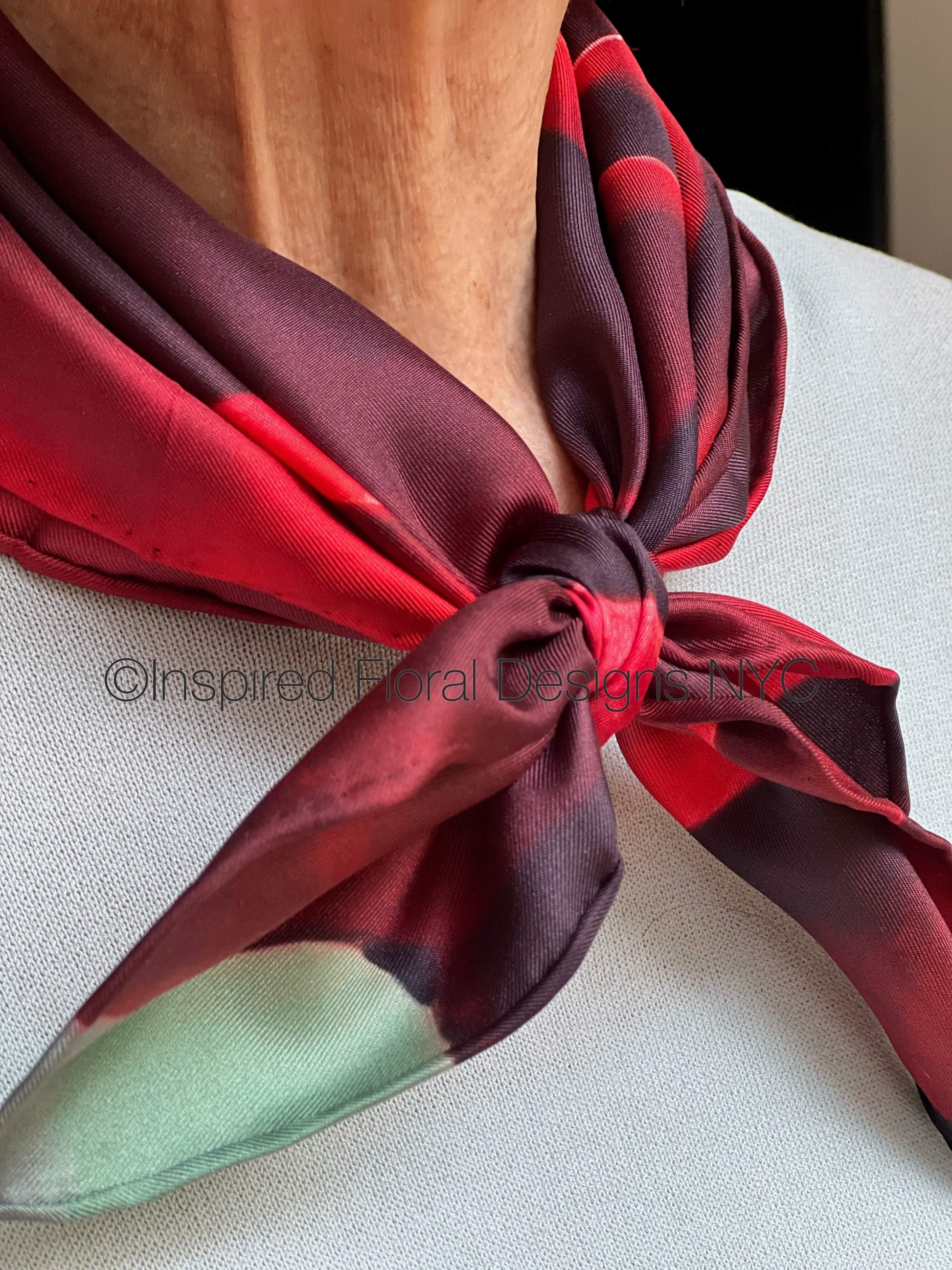 A close-up of a silk scarf tied around a person's neck. The scarf features a color-blocked pattern with dark burgundy, bright red, and mint green sections by Inspired Floral Designs NYC LLC COPYRIGHT