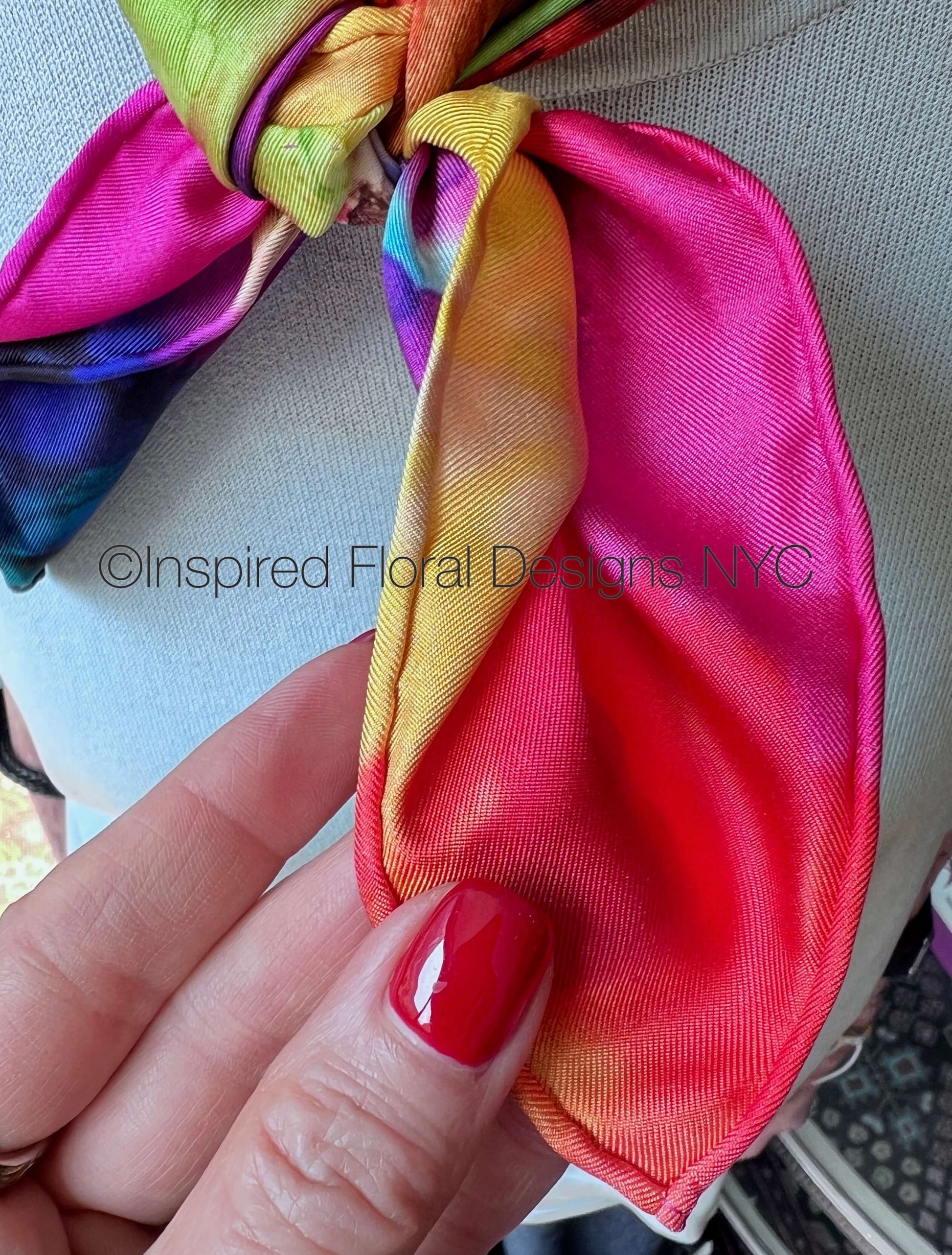 A close-up of a hand with red nail polish holding a bright pink and orange gradient silk scarf tied around a light gray fabric garment. The scarf has a satin sheen and a multicolor tie-dye pattern with yellow, green, and blue tones- silk twill scarf with hand rolled edges- inspired floral designs nyc copyright llc
