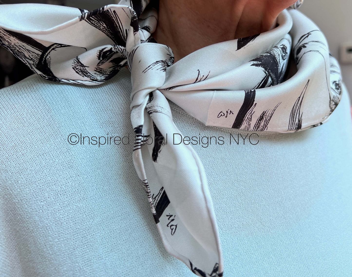 A close-up of a white silk scarf with a black brushstroke pattern of a horse head sketch tied around the neck of a person wearing a light blue knit sweater by Inspired Floral Designs NYC LLC.  A watermark 'Inspired Floral Designs NYC' is overlaid across the center. Copyright
