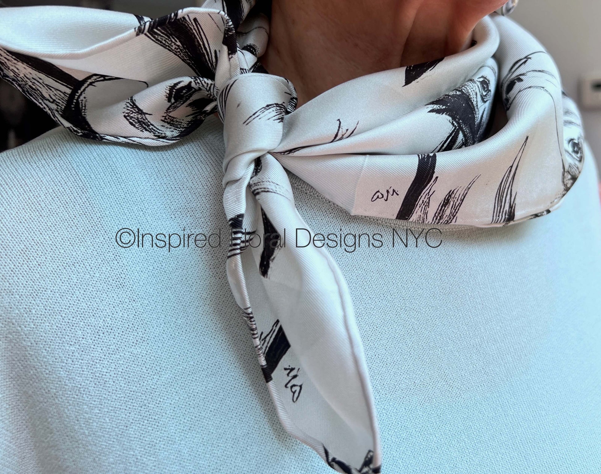 A close-up of a white silk scarf with a black brushstroke pattern of a horse head sketch tied around the neck of a person wearing a light blue knit sweater by Inspired Floral Designs NYC LLC.  A watermark 'Inspired Floral Designs NYC' is overlaid across the center. Copyright