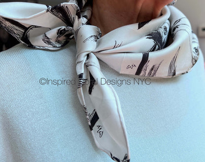 A close-up of a white silk scarf with a black brushstroke pattern of a horse head sketch tied around the neck of a person wearing a light blue knit sweater by Inspired Floral Designs NYC LLC.  A watermark 'Inspired Floral Designs NYC' is overlaid across the center. Copyright