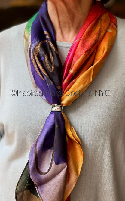 A multicolor silk scarf with purple, orange, red, and green watercolor-style patterns, tied around a person's neck with a silver ring, worn over a white shirt. By Inspired Floral Designs nyc llc copyright
