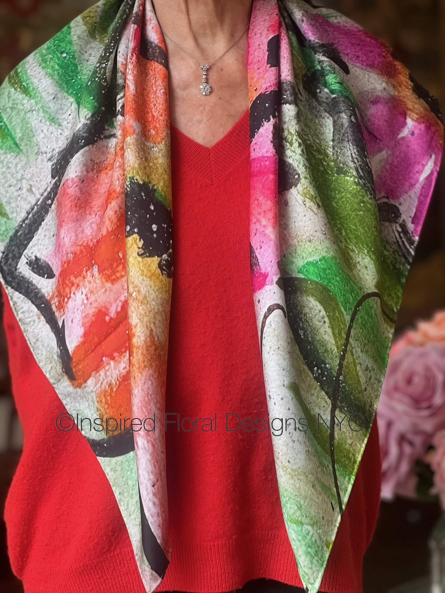 Person wearing a colorful floral scarf titled peace the edit floral art in silk over a red sweater with Inspired Floral Designs brand copyright
