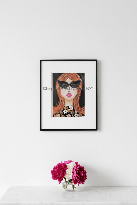 Framed artwork of a woman's face with sunglasses on a white wall, accompanied by a small vase with flowers by Inspired Floral Designs NYC LLC Copyright