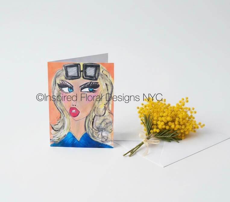 A folded greeting card featuring a stylized illustration of a woman with blonde hair, sunglasses, blue clothing, and red lips, displayed next to a small bouquet of yellow mimosa flowers on a white background by Whitney at Inspired Floral Designs NYC LLC. A watermark reading 'Inspired Floral Designs NYC' is visible across the card. Copyright