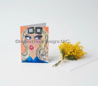 A folded greeting card featuring a stylized illustration of a woman with blonde hair, sunglasses, blue clothing, and red lips, displayed next to a small bouquet of yellow mimosa flowers on a white background by Whitney at Inspired Floral Designs NYC LLC. A watermark reading 'Inspired Floral Designs NYC' is visible across the card. Copyright