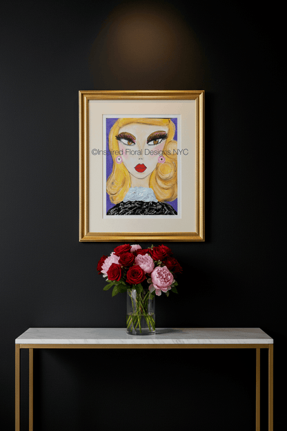 A framed art print featuring a stylized illustration of a woman with blonde hair, red lips, and a black patterned top, displayed on a dark wall above a console table with a vase of red and pink flowers by Whitney at Inspired Floral Designs NYC LLC copyright.