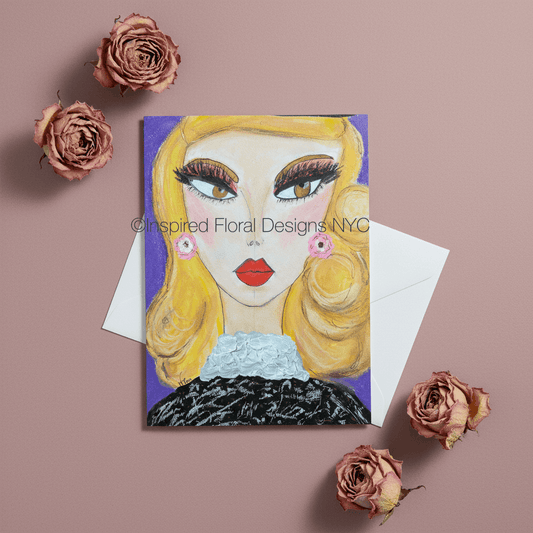 A greeting card featuring a stylized illustration of a blonde woman with bold makeup and red lips, shown with a white envelope on a dusty pink background with dried pink roses around it by Whitney at Inspired Floral Designs NYC LLC.  A watermark reading 'Inspired Floral Designs NYC' is visible across the artwork copyright.