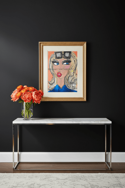 A framed art print featuring a stylized illustration of a woman with sunglasses and red lips, displayed on a dark wall above a console table with a vase of orange flowers by Whitney at Inspired Floral Designs NYC LLC copyright.