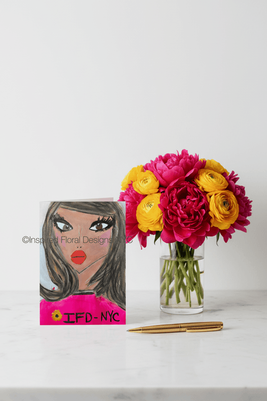 Floral arrangement in a vase next to a card with a face design on a white surface by inspired floral designs nyc llc copyright