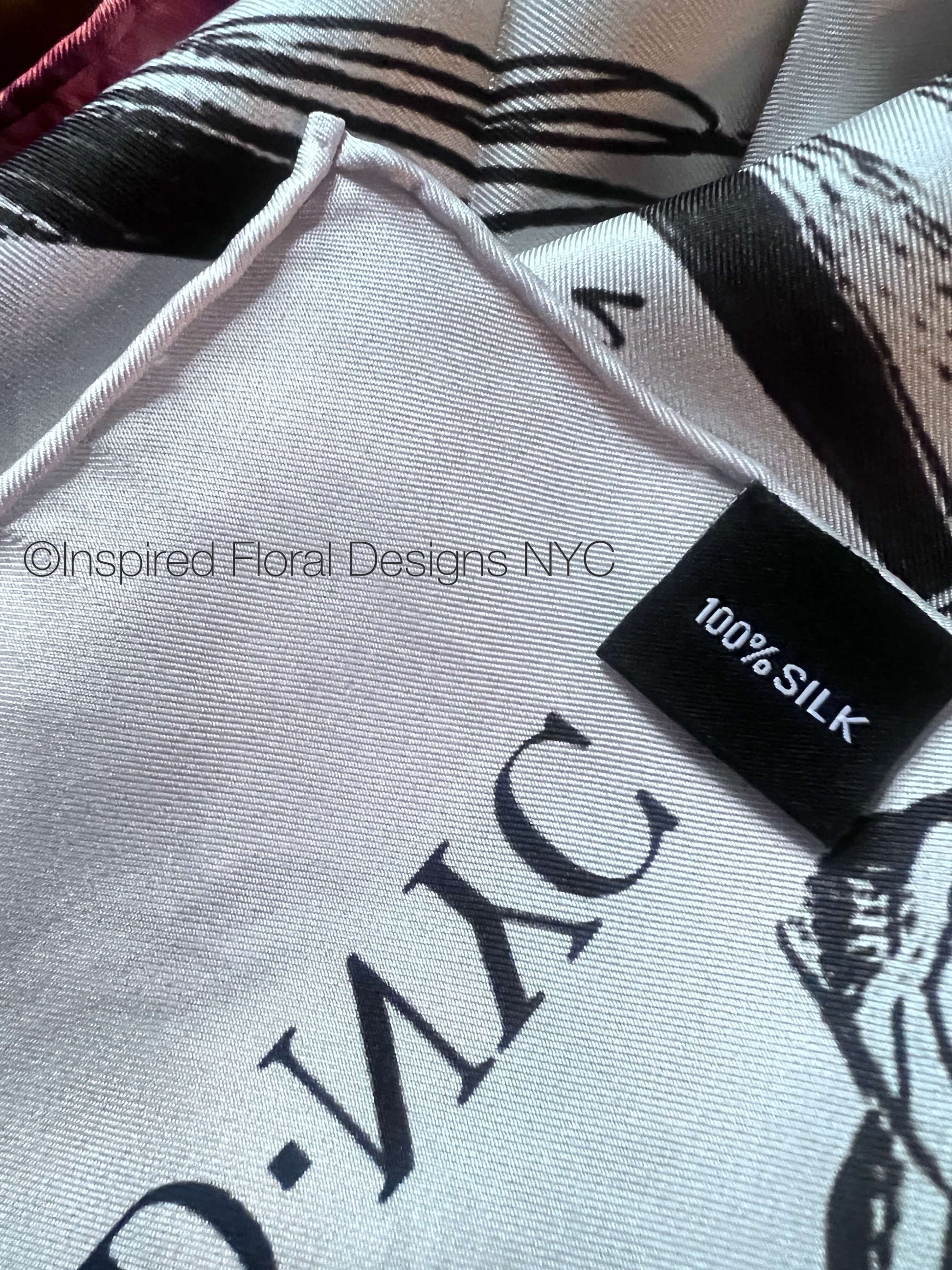 Close-up of a white silk scarf with black graphic print and a black fabric tag reading '100% SILK' and inspired floral designs nyc copyright 