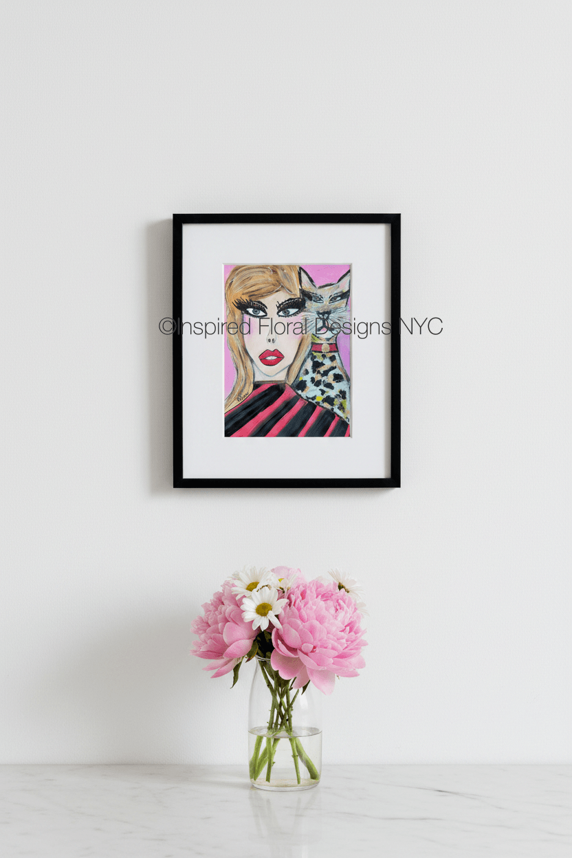 A framed art print featuring a stylized illustration of a woman with blonde hair, bold black eye makeup, red lips, and a black-and-white spotted cat on a pink background by Whitney at Inspired Floral Designs NYC LLC.  The frame is hung on a white wall above a small glass vase with pink flowers and white daisies on a light marble surface. Copyright