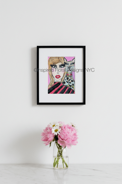 A framed art print featuring a stylized illustration of a woman with blonde hair, bold black eye makeup, red lips, and a black-and-white spotted cat on a pink background by Whitney at Inspired Floral Designs NYC LLC.  The frame is hung on a white wall above a small glass vase with pink flowers and white daisies on a light marble surface. Copyright