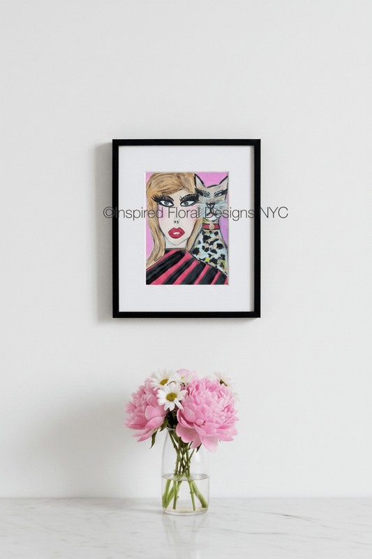 A framed art print featuring a stylized illustration of a woman with blonde hair, bold black eye makeup, red lips, and a black-and-white spotted cat on a pink background by Whitney at Inspired Floral Designs NYC LLC.  The frame is hung on a white wall above a small glass vase with pink flowers and white daisies on a light marble surface. Copyright
