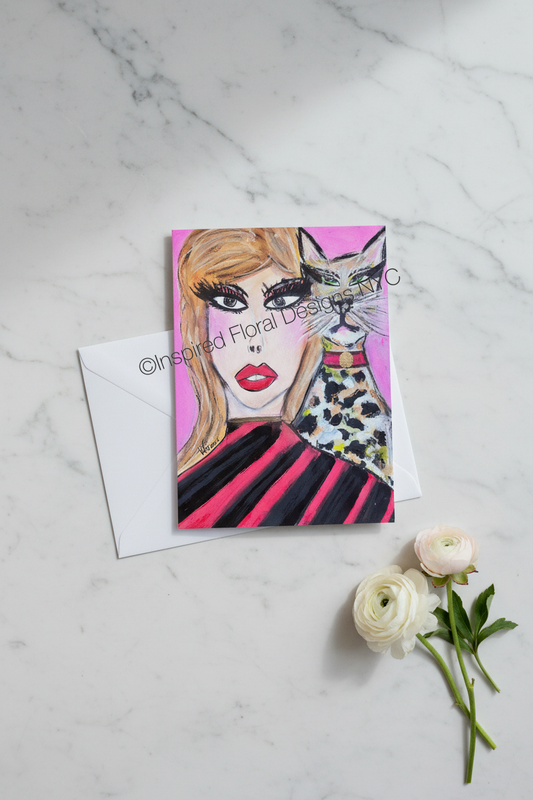 A square greeting card with a colorful illustration of a woman and a cat, placed on a white marble surface with a white envelope behind it and two white flowers at the lower right by Whitney at Inspired Floral Designs NYC LLC. The card has a visible 'Inspired Floral Designs NYC' watermark across the center.