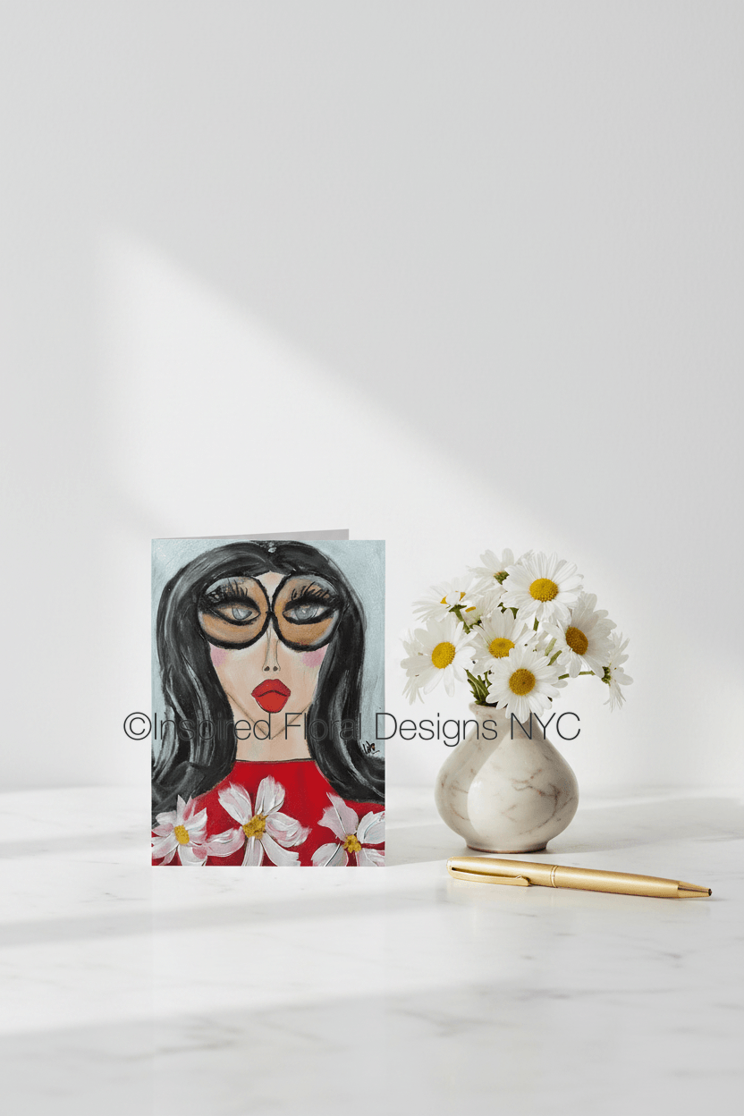 Card with a stylized face design featuring flowers, next to a vase with daisies on a white background by whitney inspired floral designs nyc llc copyright