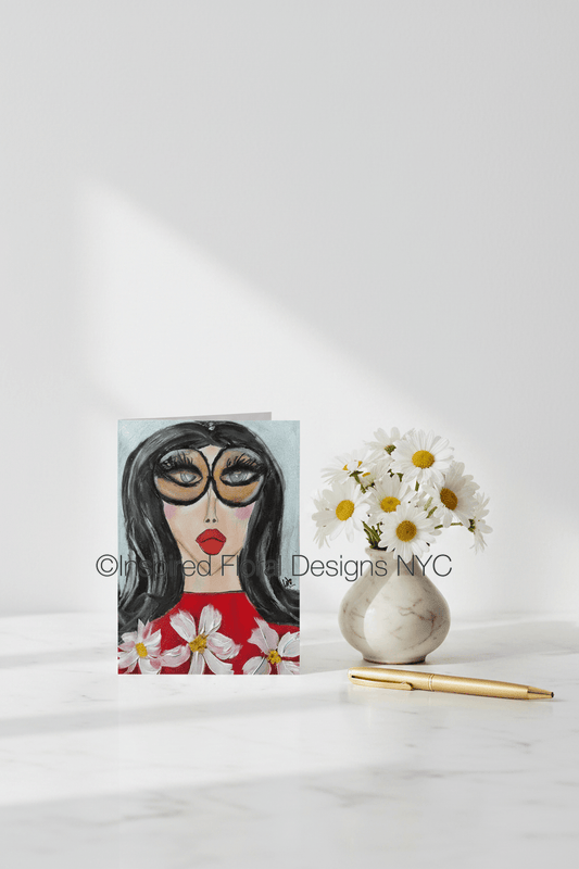 Card with a stylized face design featuring flowers, next to a vase with daisies on a white background by whitney inspired floral designs nyc llc copyright